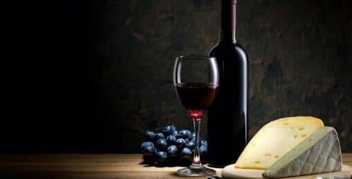 A glass of red wine, a bottle, cheese, and grapes arranged on a wooden surface in low light.