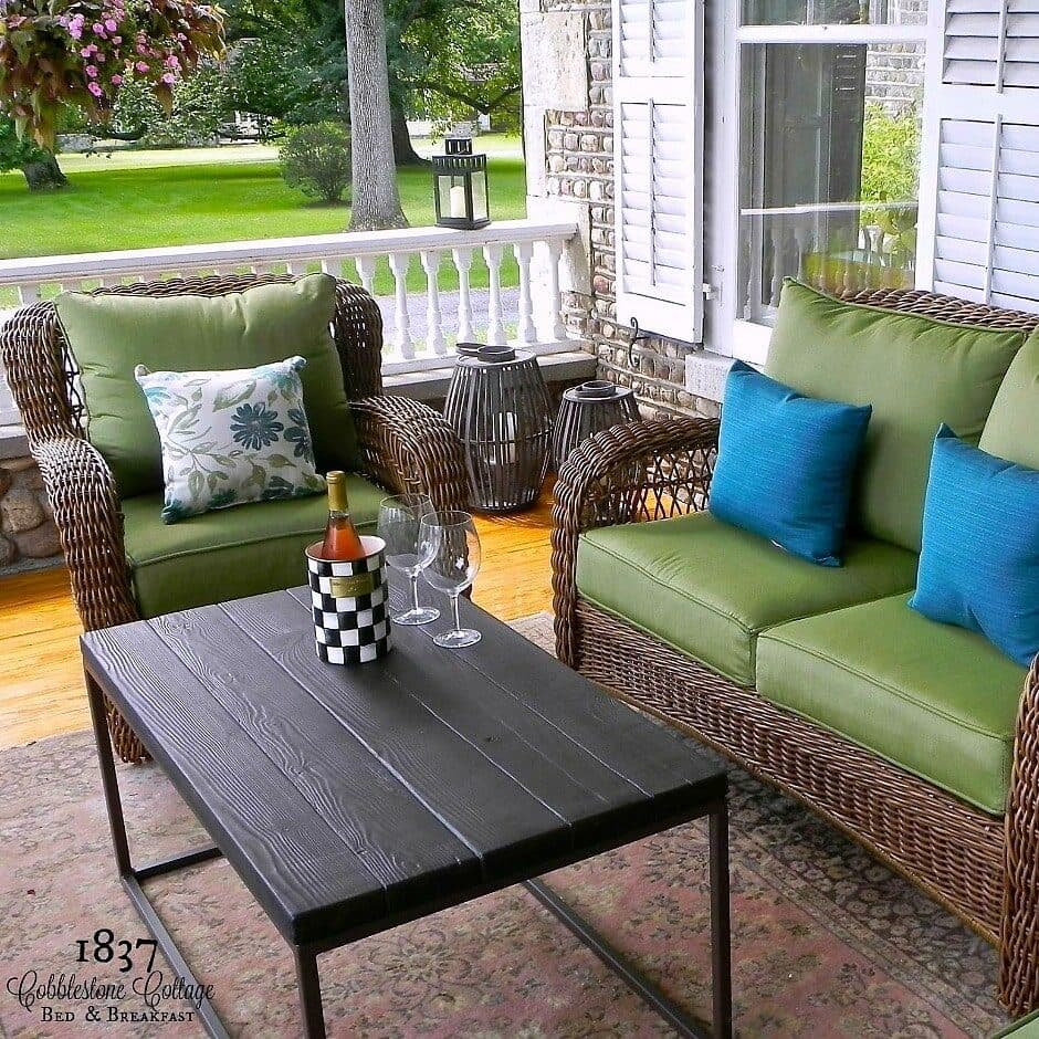 A cozy porch with green upholstered wicker chairs, a black table holding a bottle of wine and glasses, surrounded by a lush garden.