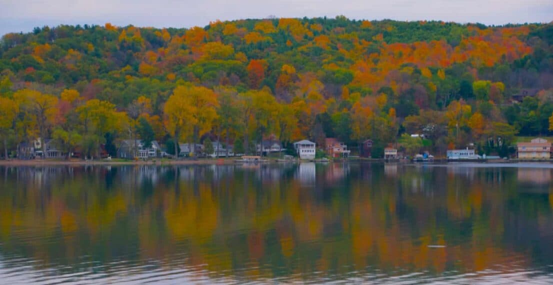 A serene lake reflects vibrant autumn foliage and a nearby shoreline of houses.