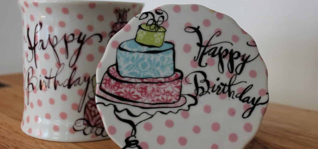 A whimsical birthday-themed ceramic mug and coaster with pink polka dots and a decorated cake illustration.