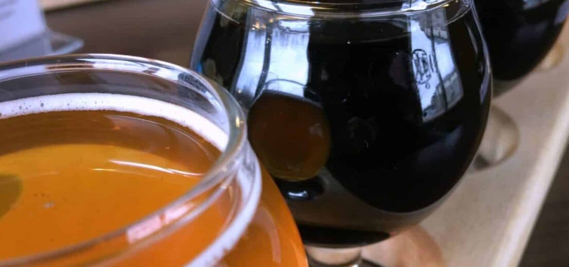 A wooden flight board holds three small glasses of assorted craft beers, featuring orange and dark black hues.