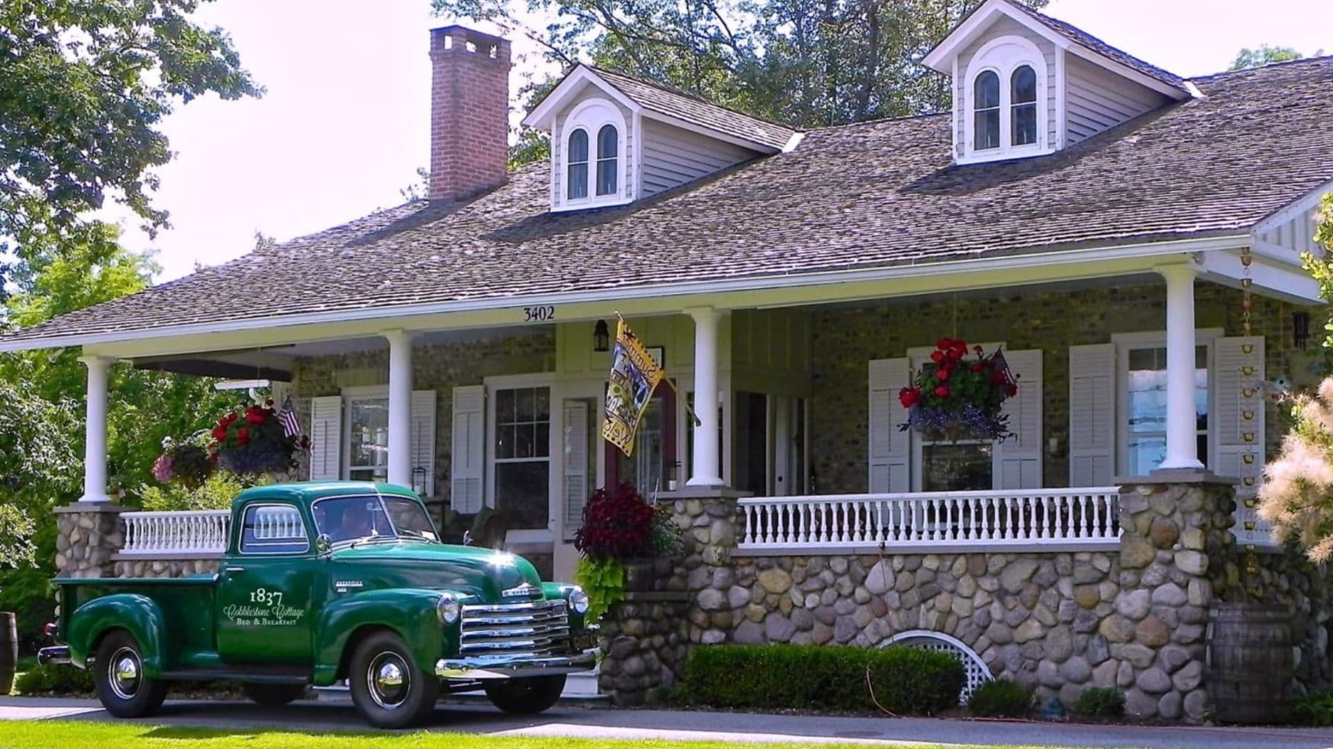 A green vintage truck parked in front of a charming stone-fronted house with a covered porch and flower boxes.